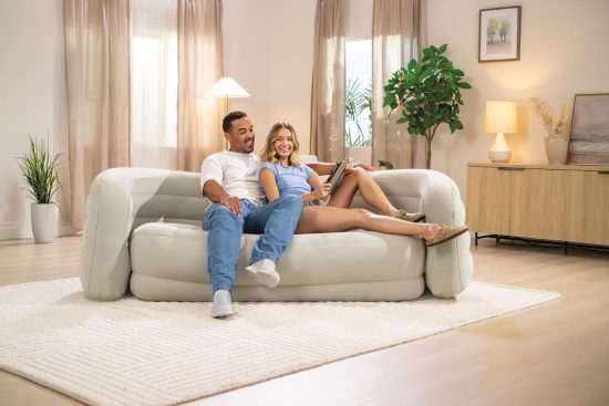 ספה מתנפחת מודרנית עם גימור פלוק (ולווט) בצבע בז' בהיר, בעיצוב מעוקל עם שלוש שכבות, מוצגת בסלון מינימליסטי מואר. על הספה כרית כחולה וכרית סרוגה לבנה. ברקע שטיח יוטה ארוג, עציץ פיקוס כינורי ושולחן קפה עם ספל ומגזין.